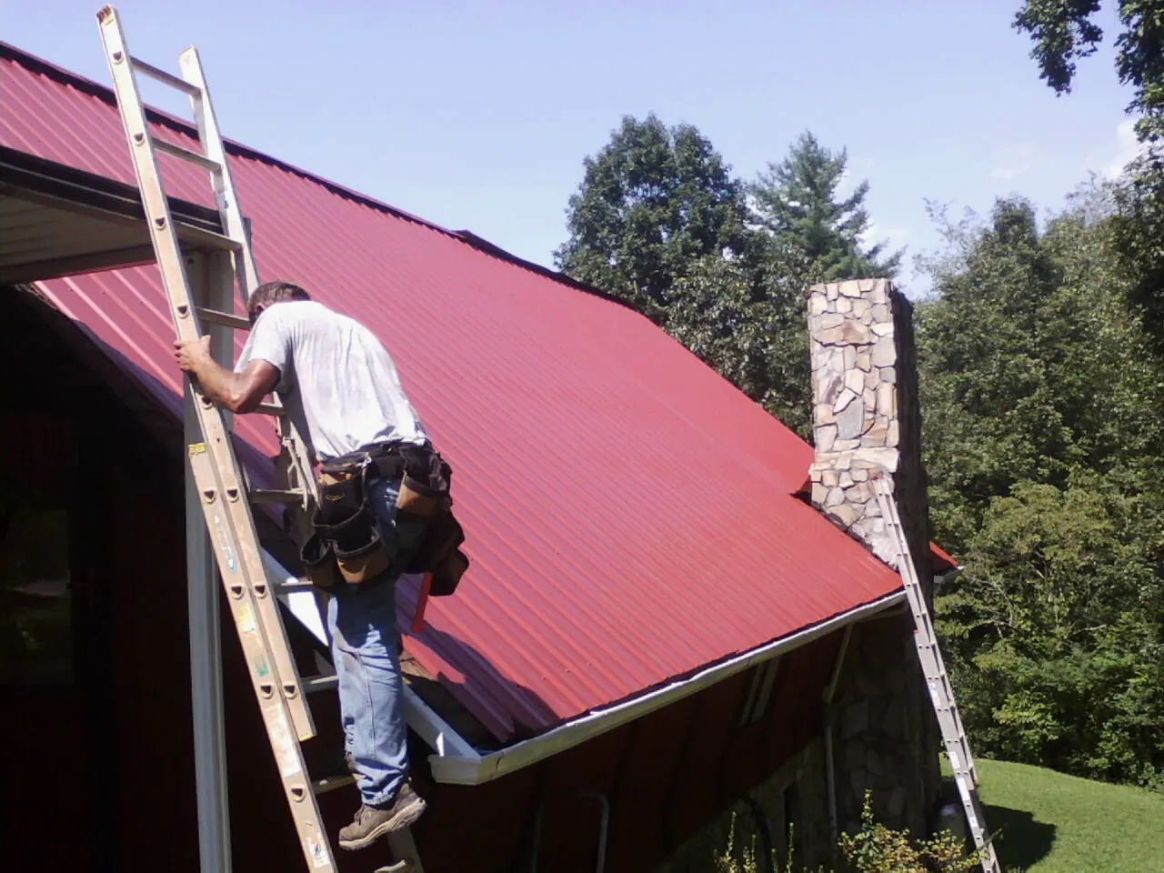 Roofer on a ladder beside a red metal roof and stone chimney — Soffit & Fascia Repair by Roofing Brothers in Selmer